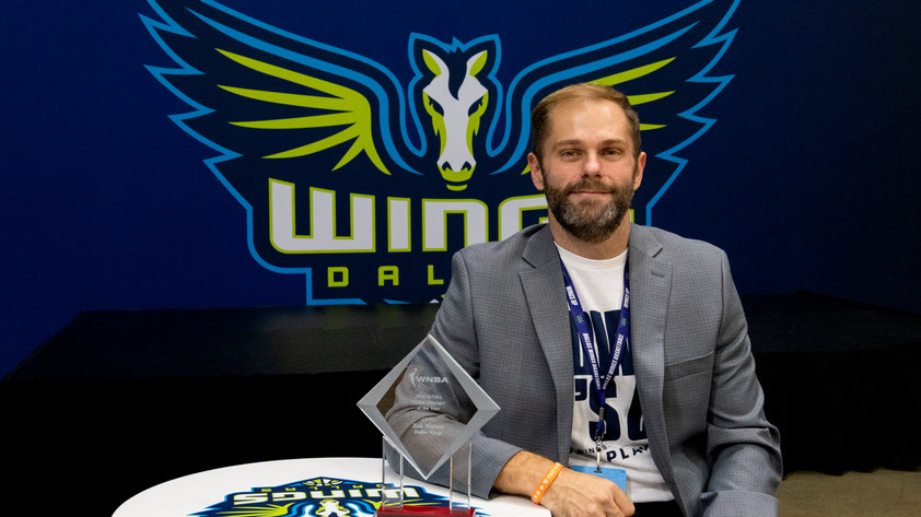 Zak Walton sitting with his 2026 W N B A Venue Manager of the Year Award. Wings Logo banner in the background